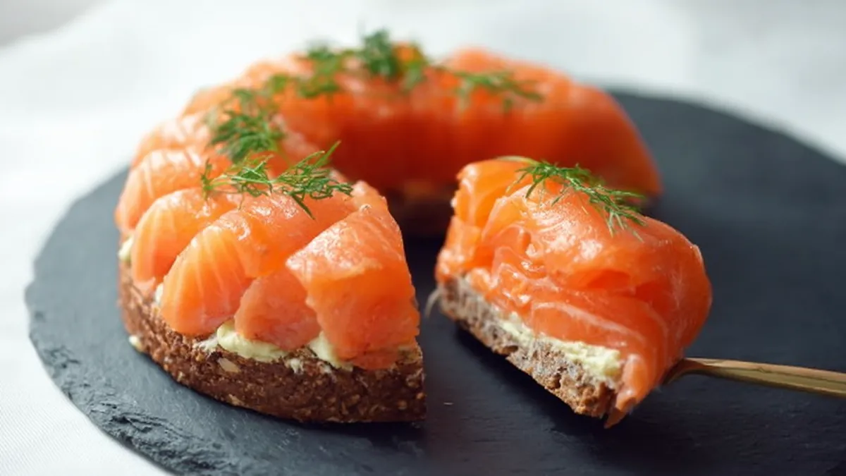Tarte apéritive au pain complet garnie de fromage frais, saumon fumé en rosace et brins d’aneth sur ardoise foncée
